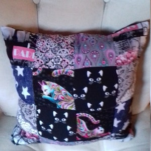Puede incluir: Una almohada de patchwork con fondo negro y una variedad de patrones coloridos, incluyendo rosa, azul y blanco. La almohada presenta un patr&oacute;n repetitivo de gatos negros con orejas y ojos blancos. La palabra "PARIS" est&aacute; impresa en rosa en la almohada.