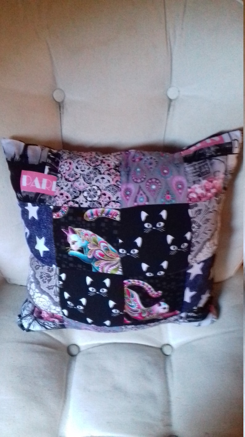 Puede incluir: Una almohada de patchwork con fondo negro y una variedad de patrones coloridos, incluyendo rosa, azul y blanco. La almohada presenta un patr&oacute;n repetitivo de gatos negros con orejas y ojos blancos. La palabra "PARIS" est&aacute; impresa en rosa en la almohada.