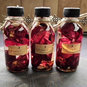 May include: Three clear glass bottles filled with red flower petals and liquid. Each bottle has a black cap, a label that reads "Aphrodite's Passion", and is adorned with twine and a decorative metal charm. The bottles are arranged on a dark surface.