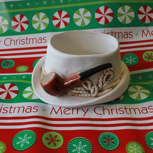 May include: A white ceramic planter shaped like a hat with a brown pipe attached to the side. The planter is decorated with a rope design and is sitting on a red and green patterned surface.