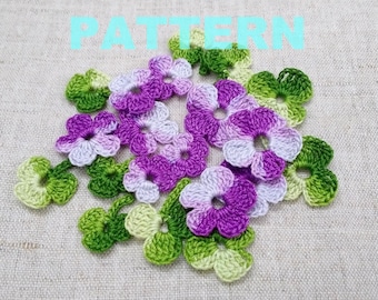 May include: A collection of crocheted flowers in shades of green, purple, and white. The flowers are arranged in a cluster on a beige fabric background.