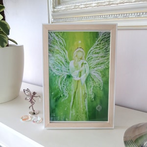 Op de afbeelding: Een ingelijst aquarel schilderij van een engel met groene vleugels en een witte jurk. De engel houdt een klein vogeltje in zijn handen. De achtergrond is een groene en witte aquarel was.