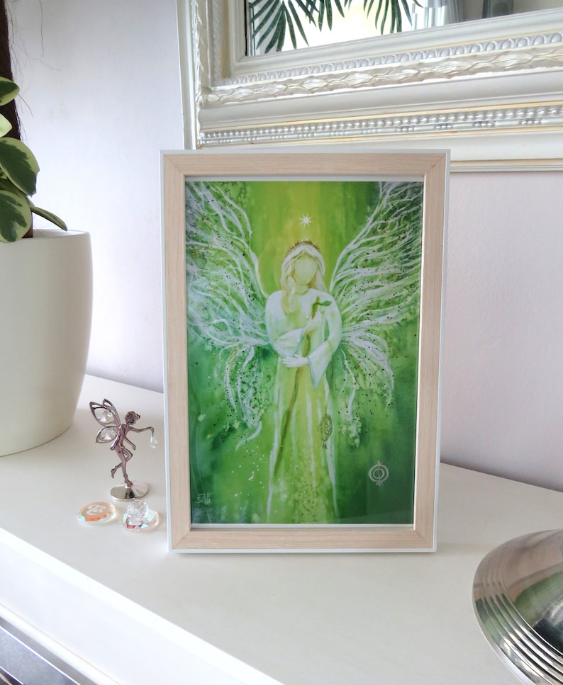 Op de afbeelding: Een ingelijst aquarel schilderij van een engel met groene vleugels en een witte jurk. De engel houdt een klein vogeltje in zijn handen. De achtergrond is een groene en witte aquarel was.