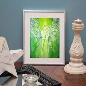Op de afbeelding: Een ingelijst aquarel schilderij van een engel met witte vleugels en een groene achtergrond. De engel houdt een staf vast en heeft een halo boven zijn hoofd.