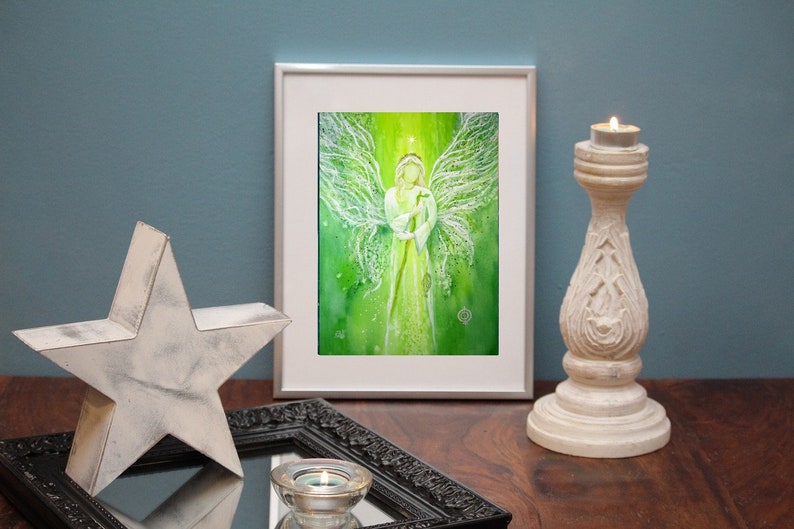 Op de afbeelding: Een ingelijst aquarel schilderij van een engel met witte vleugels en een groene achtergrond. De engel houdt een staf vast en heeft een halo boven zijn hoofd.