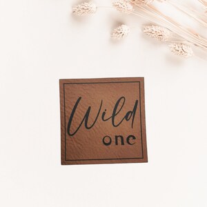 Könnte beinhalten: Eine braune Karte mit schwarzem Rand und dem Text "Wild one" in schwarzer Schrift.