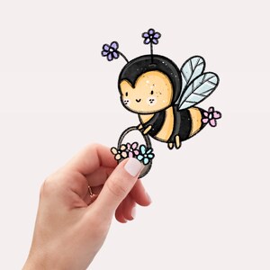 Könnte beinhalten: Eine Cartoon-Biene mit schwarzen und gelben Streifen hält einen Korb mit Blumen. Die Biene hat ein süßes Gesicht mit großen Augen und einem Lächeln. Die Blumen sind rosa, blau und gelb.
