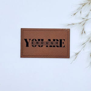 Könnte beinhalten: Ein rechteckiger brauner Lederaufnäher mit den Worten "YOU ARE ENOUGH" in schwarzen Blockbuchstaben. Der Aufnäher hat einen dunkelbraunen Rand und ist vor einem weißen Hintergrund mit federartigen Akzenten platziert.