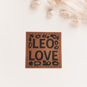 Könnte beinhalten: Eine braune Karte mit einem gesteppten Rand und schwarzem Text, der "LEO LOVE" lautet, umgeben von schwarzen abstrakten Formen.