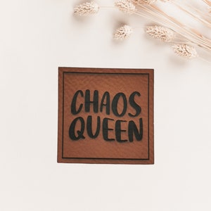 Könnte beinhalten: Ein quadratisches, braunes Holzschild mit den Worten "CHAOS QUEEN" in fetten, schwarzen Buchstaben. Das Schild hat einen dunkelbraunen Rand und ist vor einem weißen Hintergrund platziert, mit getrockneten Weizenähren in der oberen rechten Ecke.