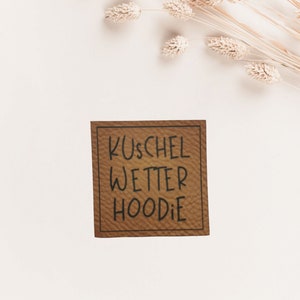 Könnte beinhalten: Ein braunes Leder-Etikett mit dem Text "Kuschel Wetter Hoodie" in schwarzer Schrift.