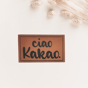 Könnte beinhalten: Ein braunes rechteckiges Schild mit dem Text "ciao Kakao." in Schwarz gedruckt.