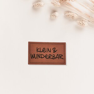 Könnte beinhalten: Ein braunes Leder-Etikett mit dem Text "klein & wunderbar" in schwarzer Schrift.