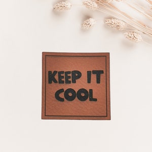 Könnte beinhalten: Ein braunes Lederstück mit schwarzem Text, der "KEEP IT COOL" auf einem weißen Hintergrund mit getrocknetem beigefarbenem Gras lautet.