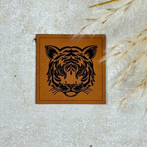 Könnte beinhalten: Ein quadratischer, brauner Lederaufnäher mit einem schwarzen Tigergesicht-Design. Das Tigergesicht ist mittig platziert, mit detaillierten Linien, die seine Merkmale definieren. Der Aufnäher ist wahrscheinlich zum Nähen oder Basteln.