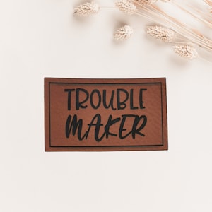 Könnte beinhalten: Ein rechteckiger brauner Lederaufnäher mit den Worten "TROUBLE MAKER" in fetter schwarzer Schrift. Der Aufnäher hat einen dünnen schwarzen Rand. Getrocknete Weizenähren sind im Hintergrund.
