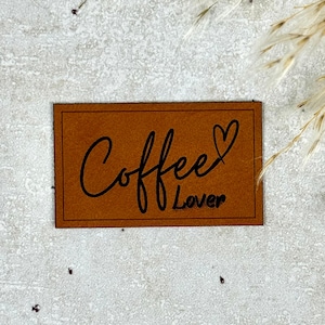 Könnte beinhalten: Ein rechteckiger, brauner Lederaufnäher mit den Worten "Coffee Lover" in schwarzer Schrift. Das Wort "Coffee" ist in Schreibschrift geschrieben, und ein Herz ersetzt den Buchstaben "e" in "Lover".