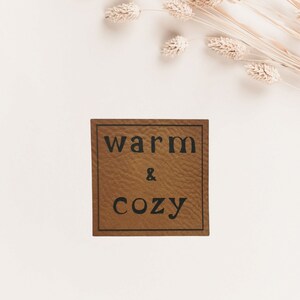 Könnte beinhalten: Ein braunes Leder-Etikett mit dem Text "warm & cozy" in schwarzen Buchstaben.