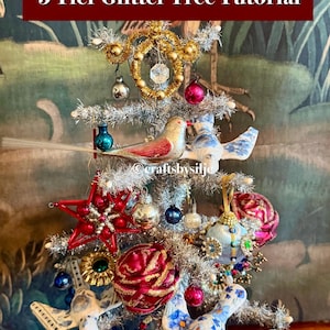 May include: A silver tinsel tree decorated with various ornaments including bird figures, red stars, and colorful baubles. The tree is topped with a wire star and features the text 5 Tier Glitter Tree Tutorial and craftsbysilje.