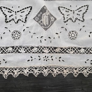 May include: A white fabric runner featuring intricate cutwork embroidery of butterflies and geometric diamond shapes. The textile includes circular motifs, delicate vine patterns, and a scalloped lace border with floral details along the bottom edge.