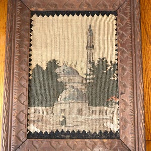 May include: A framed tapestry depicting a mosque with a tall minaret and surrounding trees. The tapestry is in shades of beige, green, and gray, set within a carved wooden frame.