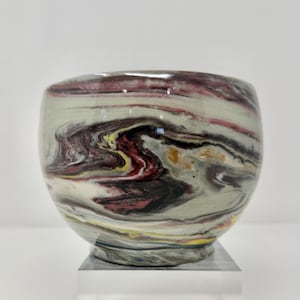 May include: A ceramic bowl with a marbled design. The bowl features swirling patterns of burgundy, black, yellow, and white against a pale green background. The bowl is round with a slightly flared rim and sits on a clear base.
