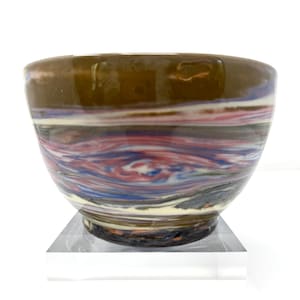 May include: A handcrafted ceramic bowl with a unique marbled design. The bowl features a dark brown rim and a swirling pattern of red, blue, white, and gray. The bowl is set on a clear acrylic stand, showcasing its artistic design.