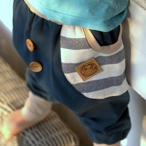 Può includere: Un paio di pantaloni a righe blu e bianche con una tasca a toppa sul lato destro. La tasca ha un'etichetta marrone con un logo di coniglio bianco e il testo "Handmade".