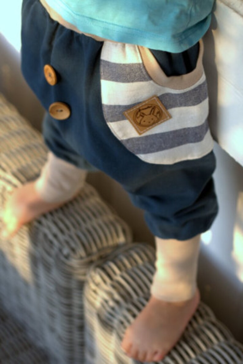 K&ouml;nnte beinhalten: Ein Kind tr&auml;gt eine blaue und wei&szlig;e gestreifte Hose mit einem braunen Flicken auf der Tasche. Die Hose hat zwei braune Kn&ouml;pfe vorne.