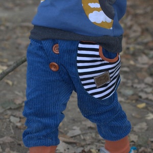 Può includere: Pantaloni di velluto a coste blu con grandi bottoni marroni e una tasca a righe. La tasca ha una toppa in pelle marrone con un logo. I pantaloni fanno parte di un outfit per bambini.