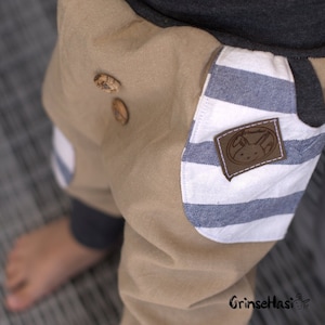 K&ouml;nnte beinhalten: Beigefarbene Hose mit blau-wei&szlig; gestreiftem Stoff auf der Tasche. Die Tasche hat einen braunen Patch mit einem wei&szlig;en Hasenlogo und dem Text "Grinsehasi".