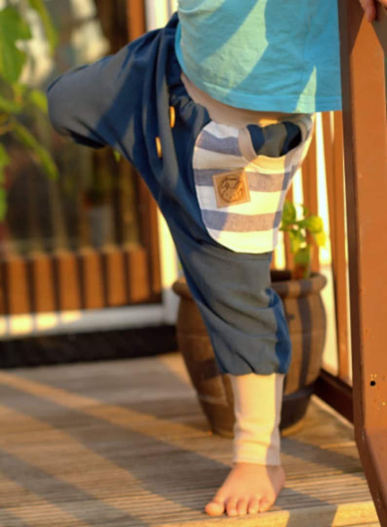 K&ouml;nnte beinhalten: Ein Kind tr&auml;gt eine blaue und wei&szlig;e gestreifte Hose mit einem Patch auf der Vorderseite. Die Hose ist locker geschnitten und hat einen Kordelzugbund. Das Kind steht auf einem Holzboden.