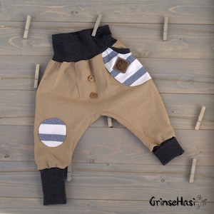 K&ouml;nnte beinhalten: Beigefarbene Babyhose mit einer gestreiften Tasche und einem gestreiften Flicken am Knie. Die Hose hat einen dunkelgrauen Bund und B&uuml;ndchen. Die Hose hat zwei braune Kn&ouml;pfe vorne.