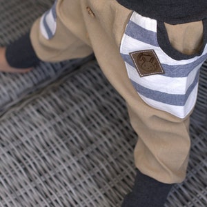 K&ouml;nnte beinhalten: Beigefarbene Hose mit blauen und wei&szlig;en Streifen aus Stoff. Ein Patch hat einen braunen gestickten Hasen mit dem Text "GrinseHasis" darauf.