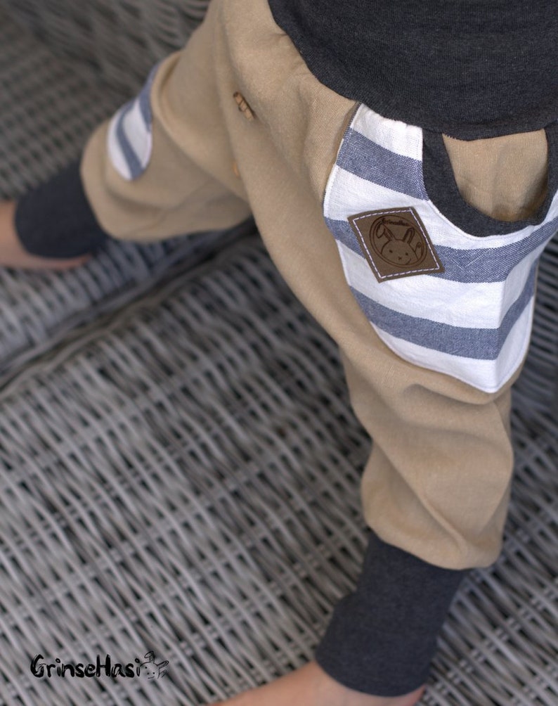 K&ouml;nnte beinhalten: Beigefarbene Hose mit blauen und wei&szlig;en Streifen aus Stoff. Ein Patch hat einen braunen gestickten Hasen mit dem Text "GrinseHasis" darauf.