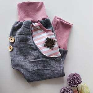 Op de afbeelding: Grijze en roze babybroek met een gestreepte zak en houten knopen. De broek heeft een roze tailleband en een bruin patch met een konijnenlogo.