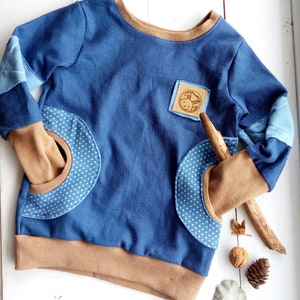Könnte beinhalten: Ein blaues Langarmshirt mit braunem Besatz und zwei blauen und weißen Polka-Dot-Taschen. Das Shirt hat einen braunen Leder-Patch mit einem kleinen Tiermotiv auf der Vorderseite.