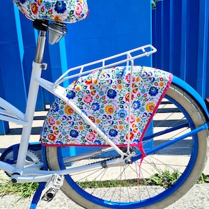 Op de afbeelding: Een witte fiets met een blauw-witte bloemenprint op de zadel en een blauw-witte bloemenprint op de tas die aan de bagagedrager is bevestigd.