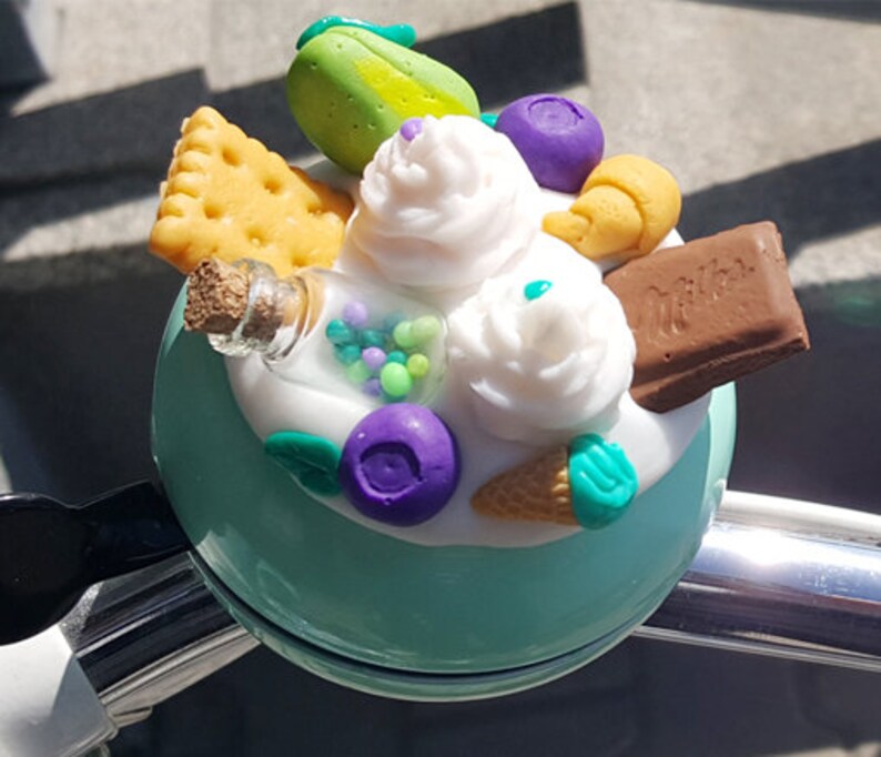 May include: A teal bicycle bell with a whimsical design featuring a white swirl, green and purple sprinkles, a chocolate bar, a green lime, a small glass bottle, and a white and blue ice cream cone.