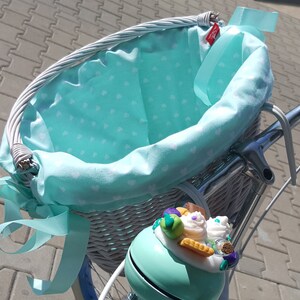 May include: White wicker bicycle basket with a light blue fabric liner with white polka dots. The basket has a light blue ribbon tied around it. A light blue bicycle bell with a decorative cupcake design is attached to the basket.