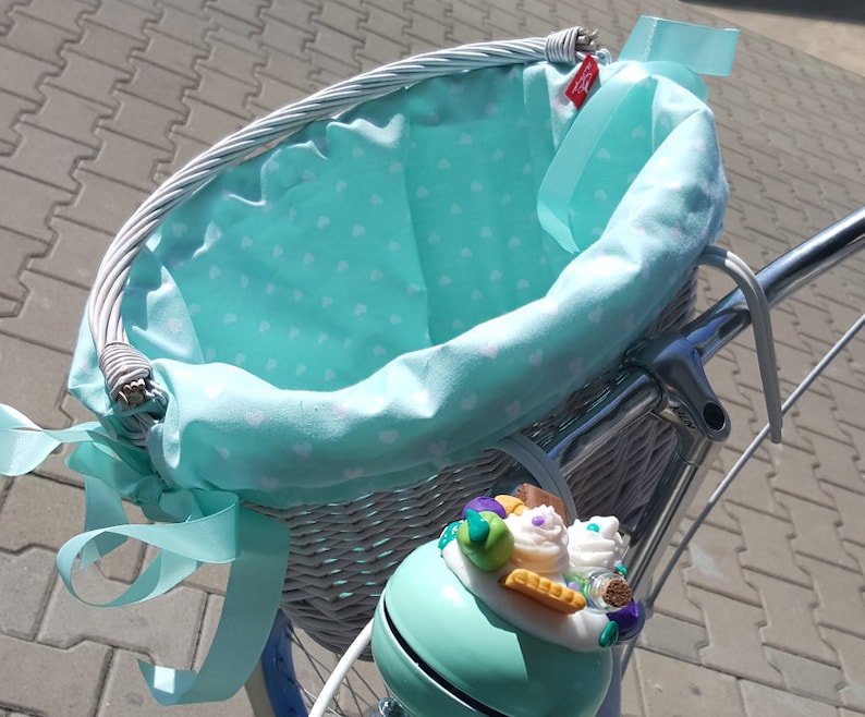 May include: White wicker bicycle basket with a light blue fabric liner with white polka dots. The basket has a light blue ribbon tied around it. A light blue bicycle bell with a decorative cupcake design is attached to the basket.