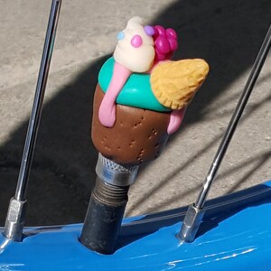 May include: A colorful ice cream cone shaped bike valve cap made of polymer clay. The cone is yellow with a brown chocolate base and topped with a white ice cream scoop with pink and blue sprinkles.