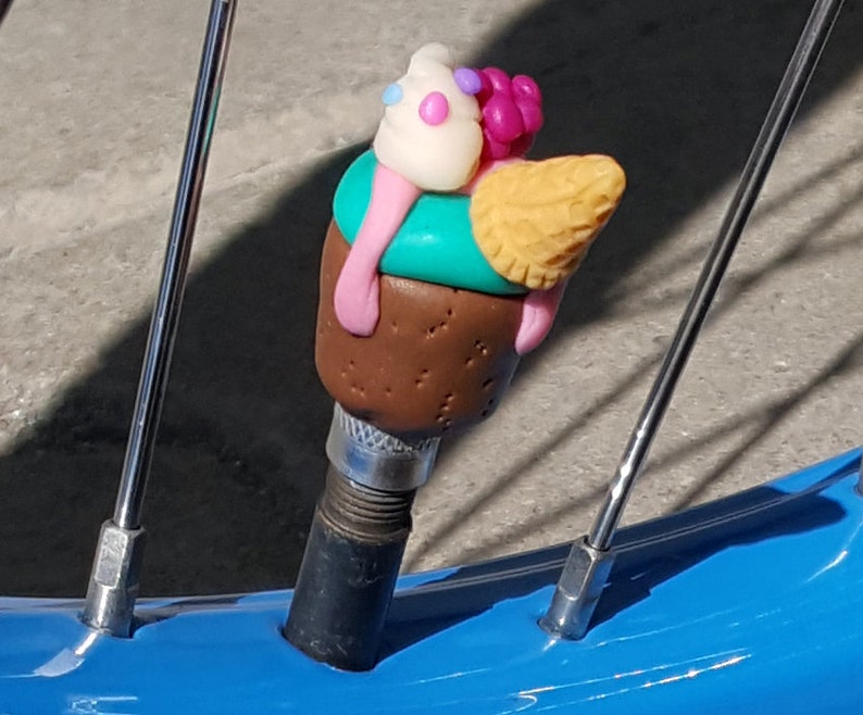 May include: A colorful ice cream cone shaped bike valve cap made of polymer clay. The cone is yellow with a brown chocolate base and topped with a white ice cream scoop with pink and blue sprinkles.