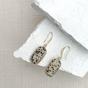 May include: A pair of dangle earrings with rectangular, speckled stone pendants in a gold-tone setting. The stones have a cream base with black spots. The earrings hang from gold-coloured ear wires.