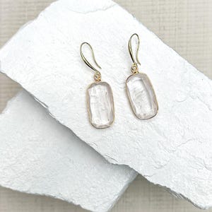 Könnte beinhalten: Ein Paar goldfarbene Ohrringe mit rechteckigen, klaren, facettierten Steinen. Die Ohrringe haben ein einfaches Haken-Design und werden auf einer weißen, strukturierten Oberfläche präsentiert. Die Steine haben einen dezenten rosa Farbton.