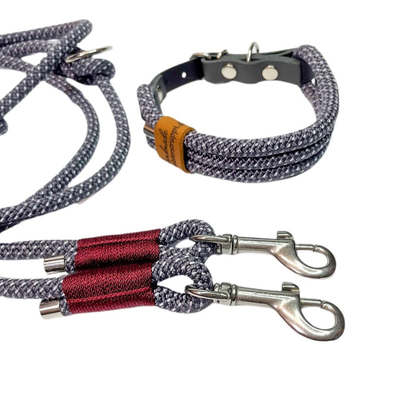 May include: A gray and white rope dog collar and leash set with a red accent on the leash. The collar has a brown leather patch with the text "Handmade with Love" on it. The leash has two silver clips.