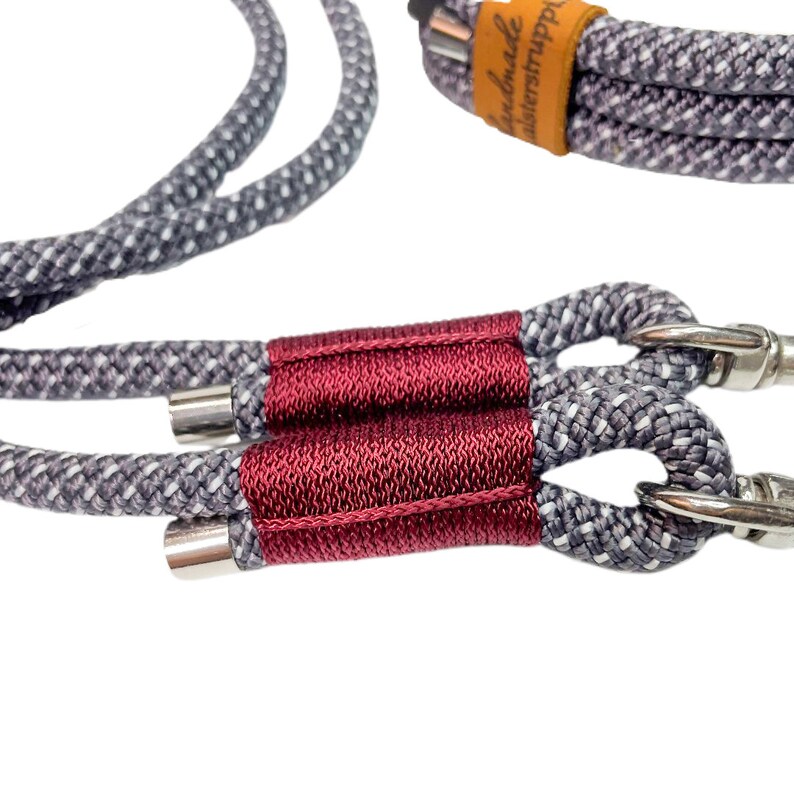 May include: A close-up of a gray and white rope dog leash with a burgundy fabric sleeve and silver metal clasps. The leash is handmade and has the text "handmade" written on a brown leather tag.