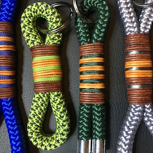 Könnte beinhalten: Vier Schlüsselanhänger aus geflochtenem Seil mit Lederakzenten. Die Schlüsselanhänger sind in Blau, Grün, Grau und Limettengrün. Jeder Schlüsselanhänger hat eine braune Lederummantelung mit orangefarbenen und gelben Streifen.