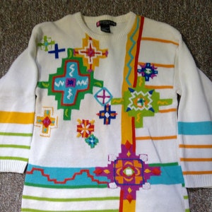 Può includere: Maglione bianco con un motivo geometrico colorato. Il motivo presenta una varietà di forme e colori, tra cui rosso, arancione, giallo, verde, blu e viola.
