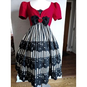 May include: A red dress with a black and white striped skirt and black lace trim. The bodice is red with a black bow and a red flower accent. The dress has short sleeves and is displayed on a mannequin.